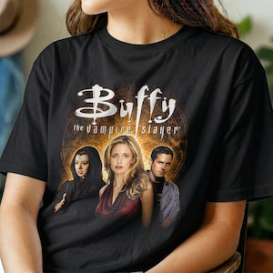 Könnte beinhalten: Ein schwarzes Kurzarm-T-Shirt mit einem Grafikdruck des Buffy the Vampire Slayer-Logos in weißem Text über drei Charakteren. Der Hintergrund der Grafik ist ein kreisförmiges, orange- und goldfarbenes Sonnenstrahlendesign mit komplexen Mustern.