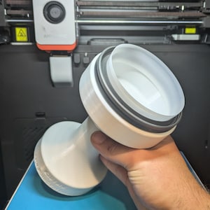 May include: A white 3D-printed object with a cylindrical shape, featuring a wide opening and a black ring around the rim. The object is held in a person's hand, with a 3D printer in the background.