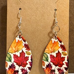 May include: Teardrop-shaped earrings featuring a colorful autumn leaf design. The earrings display a variety of fall colors, including red, orange, yellow, and green leaves, set against a white background. The earrings hang from silver-colored hooks.