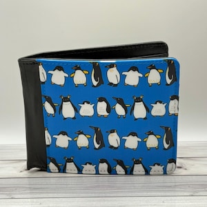 May include: A black leather bi-fold wallet with a blue panel featuring a pattern of cartoon penguins. The penguins are in various poses, with black, white, and yellow details. The wallet is closed and sits on a light-colored surface.