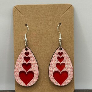May include: Teardrop-shaped earrings with a red heart design. The earrings have a white background with red text and a black border. The earrings are suspended from silver-colored hooks.
