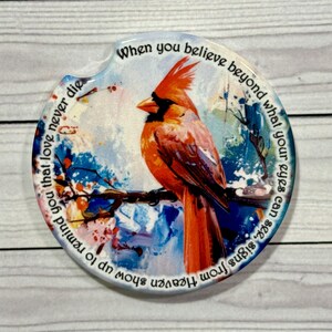May include: A round car coaster featuring a vibrant orange cardinal perched on a branch. The coaster has a watercolor background with blue, white, and red accents. Text encircles the image, reading "When you believe beyond what your eyes can see, signs from Heaven show up to remind you that love never die."