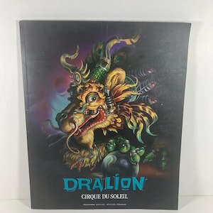 May include: A program for the Cirque du Soleil show, Dralion. The cover features a detailed illustration of a dragon-like creature with vibrant colors, including gold, green, and red, set against a dark background. The show's name is in teal letters.