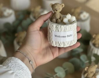 May include: A small, light-colored decorative container with a textured, tree-trunk-like exterior. It features a small teddy bear figurine and a white flower. The container has a label that reads "Welcome Baby".