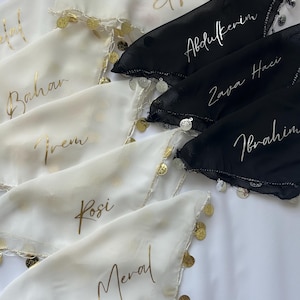 Puede incluir: Primer plano de varios pañuelos, algunos blancos y otros negros, con letras doradas. Cada pañuelo tiene un nombre diferente escrito. Los bordes están adornados con pequeñas monedas de oro y detalles plateados.