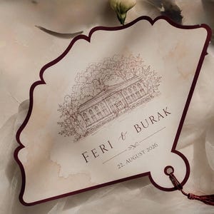 Könnte beinhalten: Eine fächerförmige Hochzeitseinladungskarte mit dunkelburgunderfarbenem Rand und passender Quaste. Das cremefarbene Papier zeigt eine detaillierte braune Strichzeichnung eines Gebäudes mit Bäumen sowie den Text FERI & BURAK und 22. AUGUST 2026.