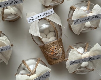 May include: Clear glass favor containers filled with white candies, decorated with twine, cream-colored bows, and tags that read "Lando Lozie." One container features a small lion cub illustration. Ideal for baby showers or special events.
