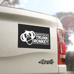 May include: A black rectangular bumper sticker with the text "PROTECTED BY TRUNK MONKEY" in white, along with a white cartoon monkey face. The sticker is affixed to the rear of a white vehicle.