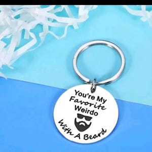 May include: A silver keyring with a circular pendant. The pendant features the text "You're My Favorite Weirdo With A Beard" in black, and a graphic of a beard and sunglasses. The keyring is set against a blue and white backdrop.