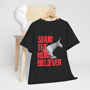 Może przedstawiać: Czarny t-shirt z krótkim rękawem z grafiką białego jednorożca i czerwonym, postarzanym napisem "SHUN THE NON-BELIEVER" z przodu. Koszulka jest wyeksponowana na drewnianym wieszaku obok gładkiej białej czapki z daszkiem.