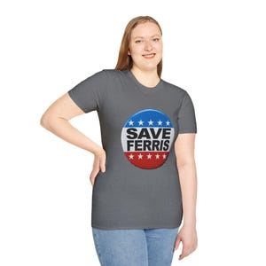 May include: A gray t-shirt featuring a circular graphic with the text "SAVE FERRIS". The graphic is designed with a blue top section with white stars, a white center with black text, and a red bottom section with white stars. The shirt is worn by a person.