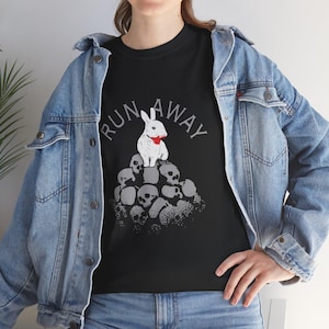 May include: Black t-shirt with a graphic design featuring a white rabbit with a red stain, perched atop a pile of skulls. The words "RUN AWAY" are arched above the rabbit in gray lettering. The shirt is made of a soft material.