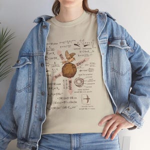 May include: Beige t-shirt featuring a detailed graphic of a bird, a globe, and scientific equations. The design includes the words "Lift", "Drag", "Thrust", and "Weight". The shirt is made of a soft material.