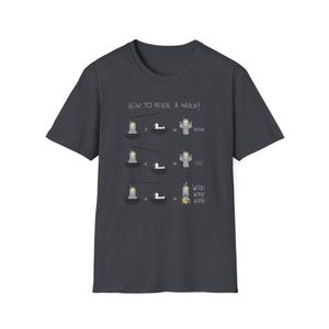 May include: Charcoal grey t-shirt featuring the phrase "HOW TO REVEAL A WITCH!" and a cartoon illustration. The design shows a witch-revealing process with a duck and a cauldron. A casual, everyday wear item.