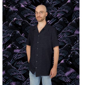 May include: A dark blue button-down shirt with a small, repeating pattern. The shirt is worn by a person standing in front of a background of black ravens with purple accents. The shirt has short sleeves and a classic collar.