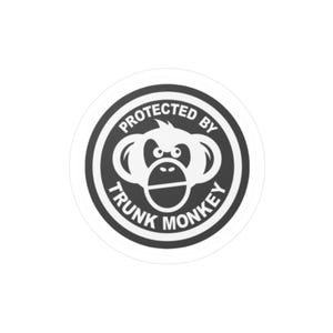 May include: A circular sticker with a white border and a black and white design. The design features a monkey face in the center, surrounded by the words "PROTECTED BY TRUNK MONKEY" in a circular pattern.