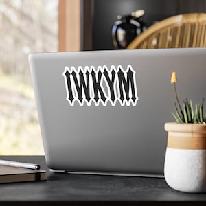 Puede incluir: Una pegatina blanca con el texto negro "IWKYM" en una fuente gótica está adherida a la parte posterior de un portátil gris. El portátil está sobre una superficie negra, con una pequeña planta en maceta y un cuaderno visibles.
