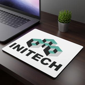 May include: White rectangular mousepad with the word "INITECH" in bold black letters. The mousepad features a geometric logo in teal and black. A laptop and small potted plant are in the background.