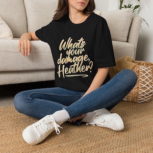 May include: A black short-sleeved t-shirt featuring the text "What's your damage, Heather?" printed in a light yellow, cursive-style font on the front. The shirt is paired with blue denim jeans and white trainers, creating a casual ensemble.