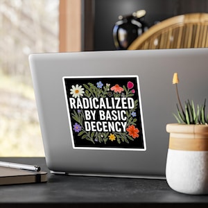 Könnte beinhalten: Ein schwarzer Aufkleber mit dem weißen Text "RADICALIZED BY BASIC DECENCY", umgeben von bunten Blumen. Der Aufkleber befindet sich auf einem Laptop, mit unscharfem Hintergrund.