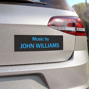 Può includere: Un magnete rettangolare nero per auto con la scritta "Music by JOHN WILLIAMS" in blu. Il magnete è attaccato alla parte posteriore di un'auto argentata, vicino al fanale.