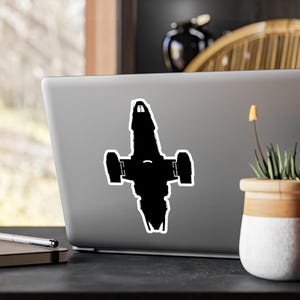 Puede incluir: Una pegatina negra con la silueta de una nave espacial con un contorno blanco. La pegatina está adherida a un portátil gris, con un fondo borroso.
