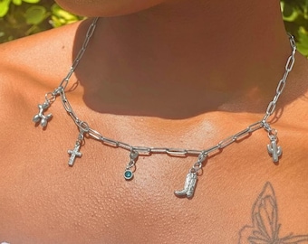 May include: Silver-toned charm necklace featuring a balloon dog, cross, teal gem, cowboy boot, and cactus pendants. The necklace has a link chain and is worn around the neck. The background is blurred green foliage.