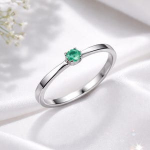 May include: A silver ring with a small, round, emerald-green gemstone set in the center. The ring band is thin and polished, reflecting light. The ring is displayed on a white, silky surface, with soft lighting.