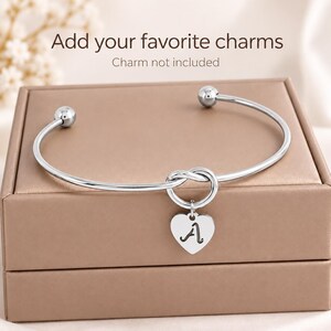May include: A silver-toned metal bangle bracelet featuring a central knot design and a heart-shaped pendant engraved with the letter A. The bracelet has spherical ends. Text above reads: Add your favorite charms. Charm not included.