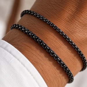 May include: Two thin, black, metallic box chain bracelets worn together. Each bracelet features a series of small, square, interlocking links with a polished, dark finish that creates a smooth, continuous, and uniform appearance.