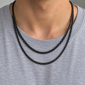 May include: A double-layered black wheat chain necklace featuring a smooth, polished finish. The two chains are of slightly different lengths, creating a tiered effect around the neck. The metal links are tightly woven in a consistent pattern.