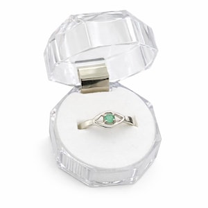 May include: A silver ring with a small, square, green gemstone set in an eye-shaped design. The ring is displayed in an open, clear, faceted plastic jewelry box with a white interior.