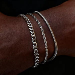 May include: Three silver bracelets of different designs are shown on a dark-skinned arm. The bracelets include a curb chain, a rope chain, and a snake chain, all in a shiny silver finish. The bracelets are stacked together.