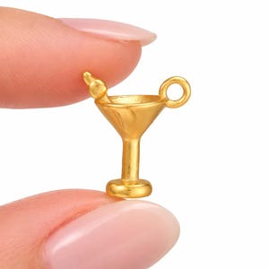 Peut inclure: Un petit charme en forme de verre à martini doré, avec une tige, une anse circulaire et une petite olive. Le charme est très détaillé et semble être en métal. Il est tenu entre deux doigts.