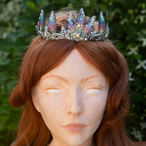 May include: A silver crown with iridescent butterfly wings and a floral design. The crown is worn on a red-haired mannequin.