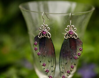 May include: A pair of silver earrings with purple iridescent wings and pink crystal accents. The earrings are hanging from a glass goblet.