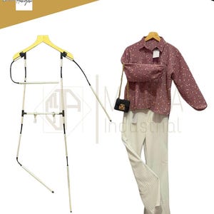 Può includere: Un'esposizione con una camicia color malva con bottoni e motivo floreale, un marsupio abbinato e pantaloni plissettati color crema. Una piccola borsa nera con una catena dorata è appesa alla camicia. È presente anche una gruccia unica.