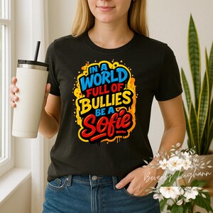 Puede incluir: Camiseta negra con el texto "IN A WORLD FULL OF BULLIES BE A Sofie" en azul, amarillo y rojo. El diseño tiene una estética de estilo graffiti. Un vaso blanco con tapa y pajita negra se sostiene en la mano izquierda.