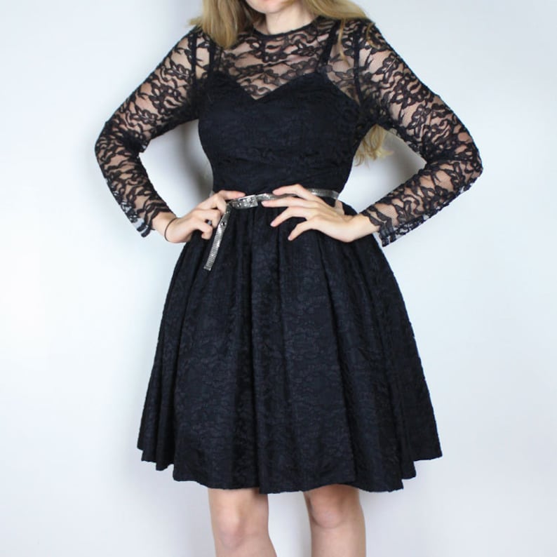 Pode incluir: Um vestido preto de renda com um corpete justo e uma saia rodada. O vestido tem mangas compridas e um cinto prateado.