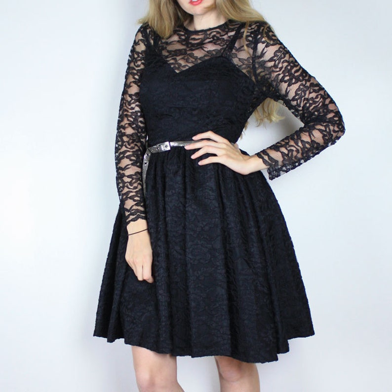 Pode incluir: Um vestido preto de renda com mangas compridas e um corpete justo. O vestido tem uma saia rodada e um cinto prateado.