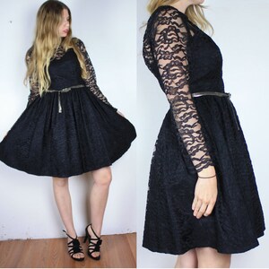 Pode incluir: Um vestido preto de renda com mangas compridas e saia rodada. O vestido tem um corpete justo e um cinto prateado.