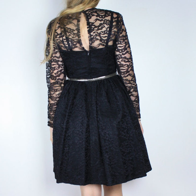 Pode incluir: Um vestido preto de renda com mangas compridas e um corpete justo. O vestido tem uma saia rodada e um cinto prateado.
