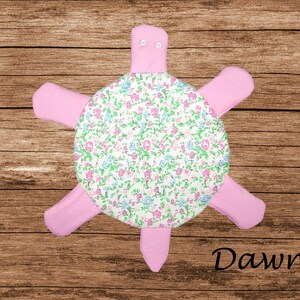 Peut inclure: Une peluche en forme de tortue avec une carapace ronde à fond blanc ornée d'un motif floral dans les tons rose, vert et bleu. La tête, les quatre pattes et la queue sont en tissu rose uni. La tête comporte deux petits boutons blancs. Texte : Dawn.