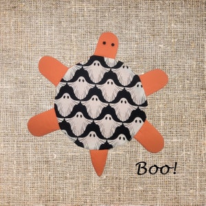 May include: A turtle-shaped fabric item with orange limbs and a head featuring two small black eyes. The central shell is covered in a repeating pattern of white ghosts on a black background. The word Boo! is printed in black in the lower right corner.