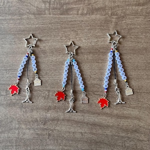May include: Three keychains with star-shaped tops, each featuring a beaded name, a red maple leaf charm, a hockey stick charm, and a house charm. The names are "SHANE ILYA", "HEATED RIVALRY", and "HOLANDER ROZANOV".