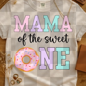 May include: White t-shirt with the text "MAMA of the sweet ONE" in pastel colors and a pink donut graphic with sprinkles. The shirt has a crew neck and short sleeves. The shirt is on a checkered background.