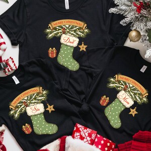 May include: Black t-shirts with Christmas-themed designs. Each shirt features a green stocking with a name, a banner that says "Merry Christmas", and a star. Festive decorations include a Santa figurine, holly, and a Christmas tree.