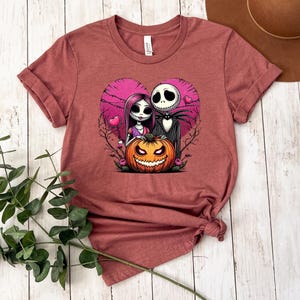 Puede incluir: Camiseta granate con un dise&ntilde;o en forma de coraz&oacute;n con Jack Skellington y Sally de Pesadilla antes de Navidad, junto con una calabaza tallada. El dise&ntilde;o es en tonos rosa, negro y naranja.