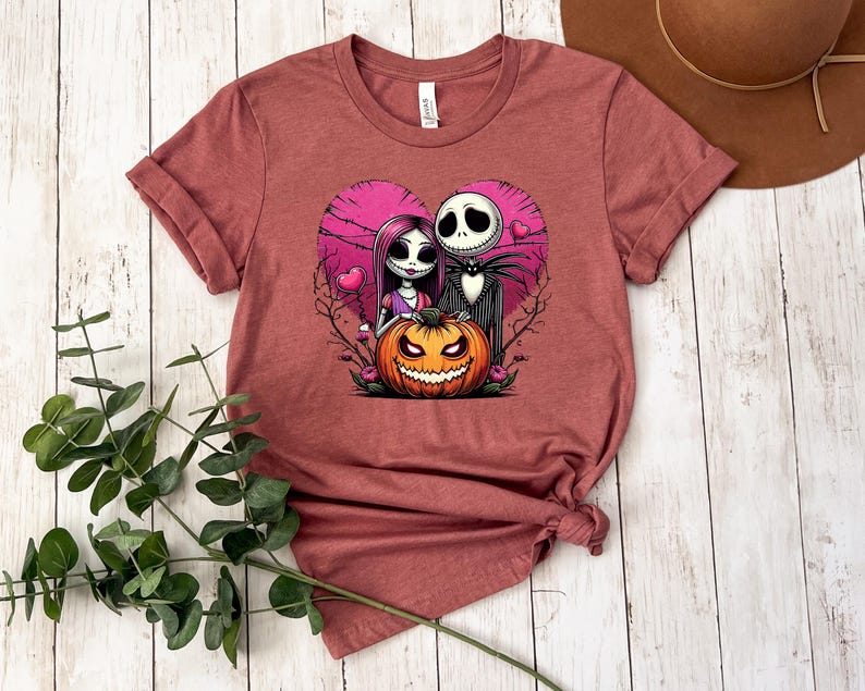 Puede incluir: Camiseta granate con un dise&ntilde;o en forma de coraz&oacute;n con Jack Skellington y Sally de Pesadilla antes de Navidad, junto con una calabaza tallada. El dise&ntilde;o es en tonos rosa, negro y naranja.