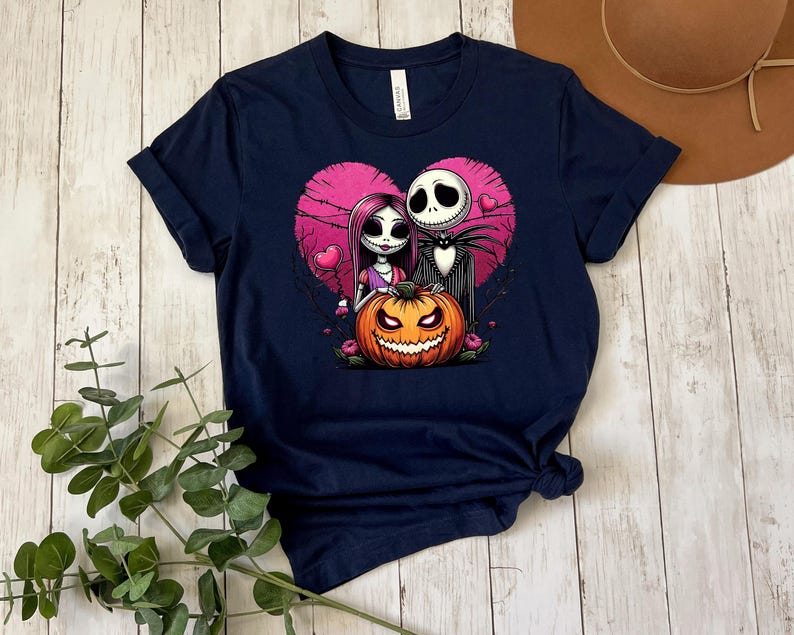 Puede incluir: Camiseta azul marino con una ilustraci&oacute;n de dibujos animados de Jack Skellington y Sally de Pesadilla antes de Navidad, con un fondo de coraz&oacute;n rosa y una calabaza. El dise&ntilde;o est&aacute; centrado en la camiseta.